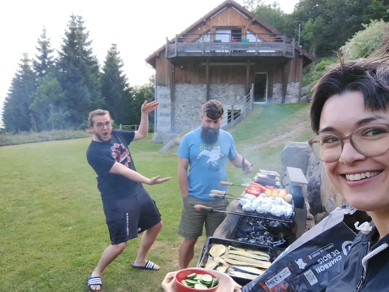 Draussen, im Hintergrund unsere Unterkunft, Andri am Grill, David daneben am Faxen machen, Sereina lacht im Vordergrund in die Kamera.