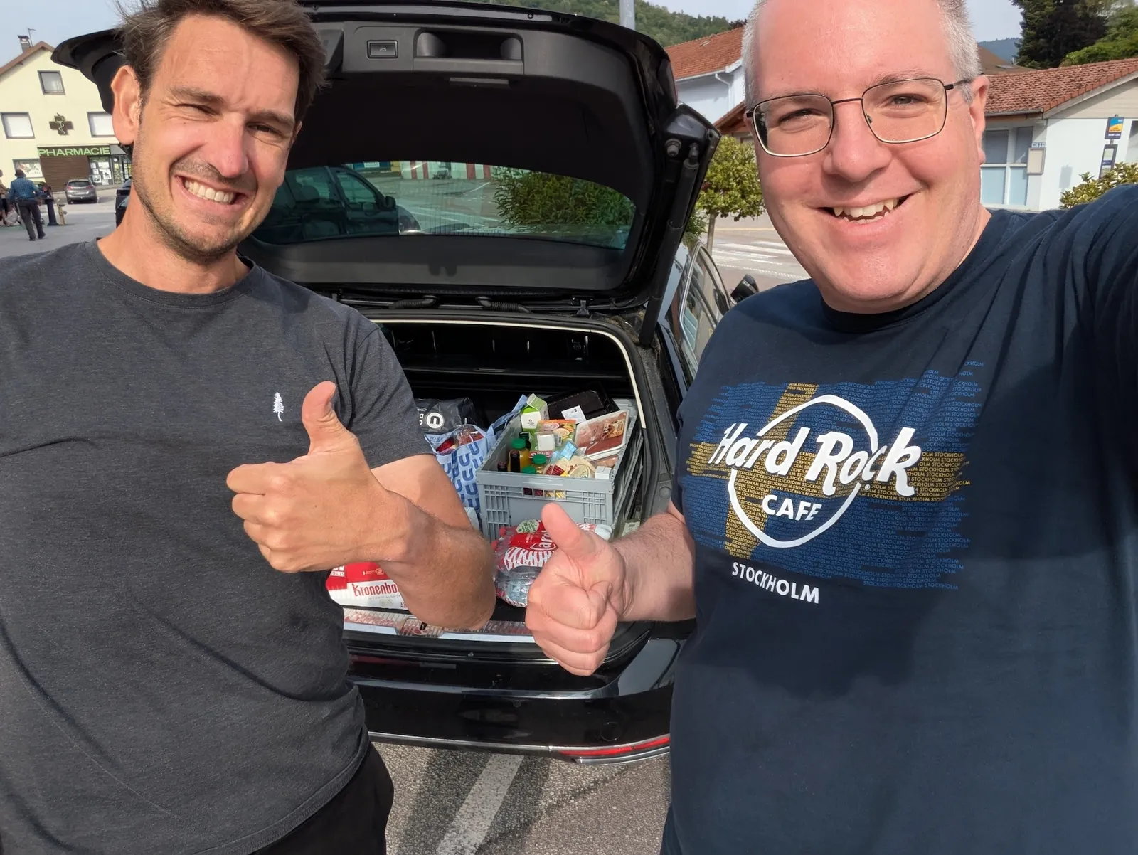 Martin und Mario stehen vor einem Auto mit offenen Kofferraum voller Essen, beide zeigen den Daumen hoch und lachen in die Kamera.