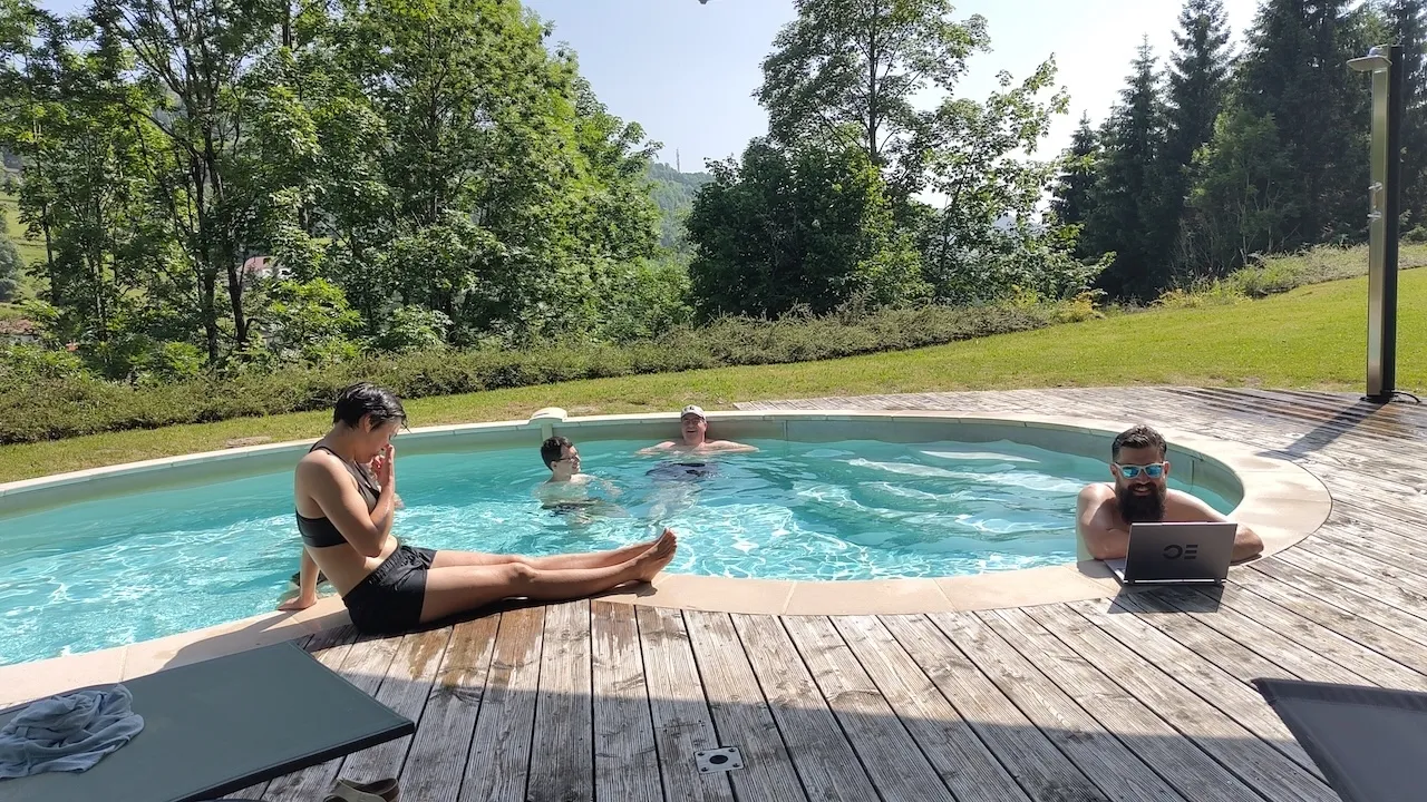 Andri steht im Wasser am Poolrand und arbeitet am Laptop, Sereina sonnt sich am Rand, Rest ist im Pool.