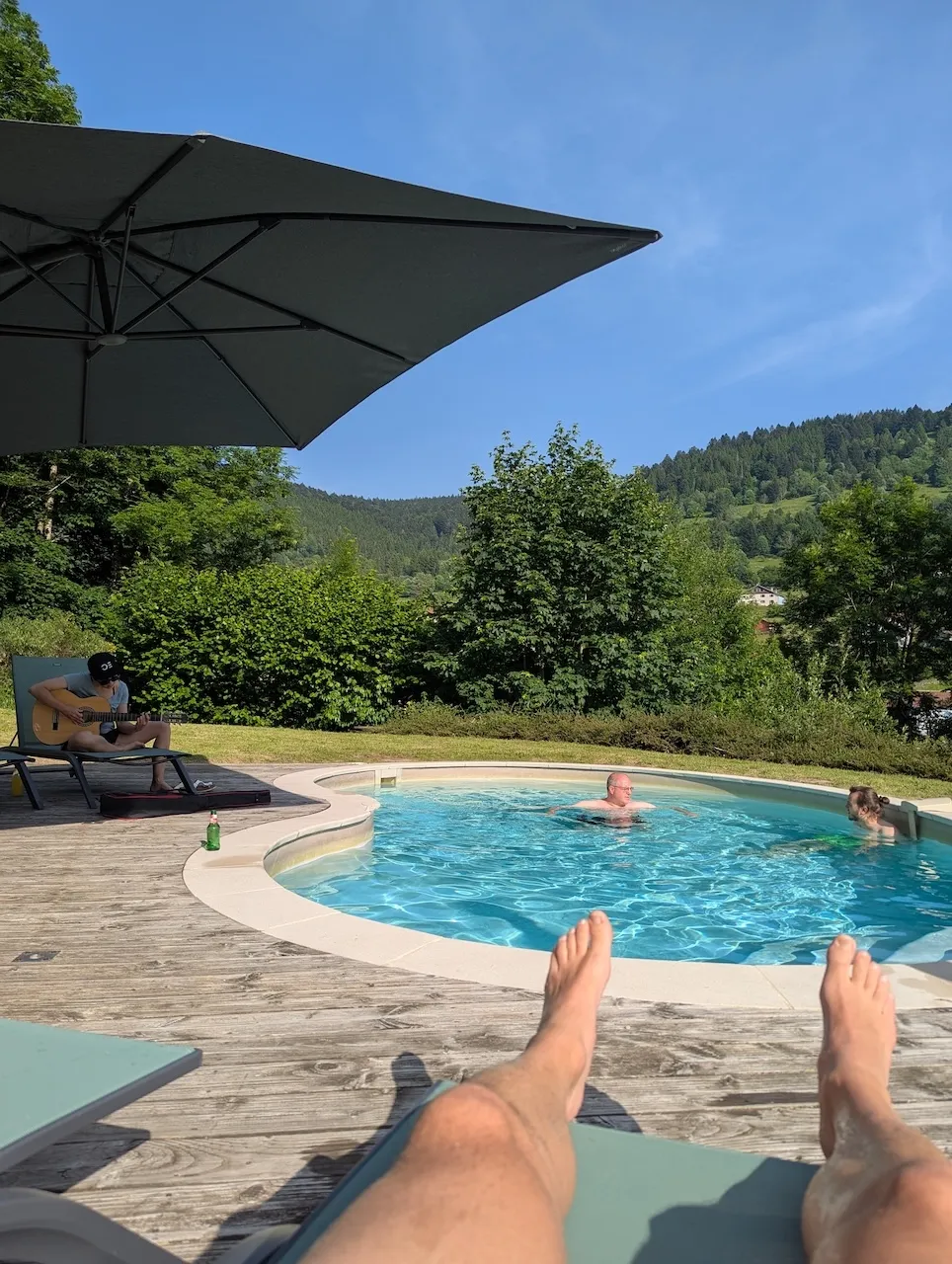 Im Vordergrund Füsse auf einem Liegestuhl, dahinter Menschen im Pool, seitlich eine Person auf einem Liegestuhl am Gitarre spielen.