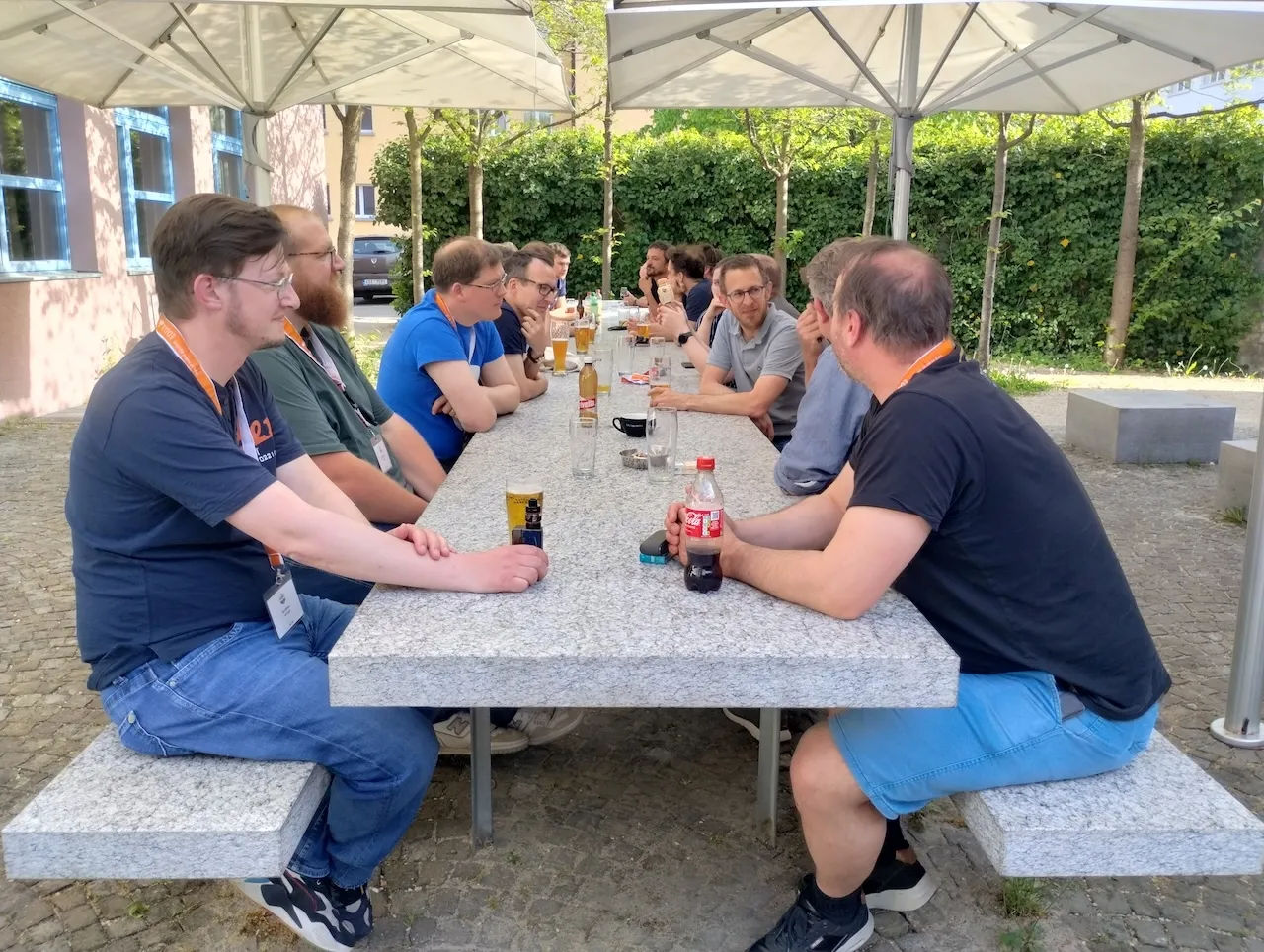 Camp-Teilnehmende an einem sehr langen Steintisch mit grossen Sonnenschirmen, fast alle haben ein kühles Bier in der Hand.